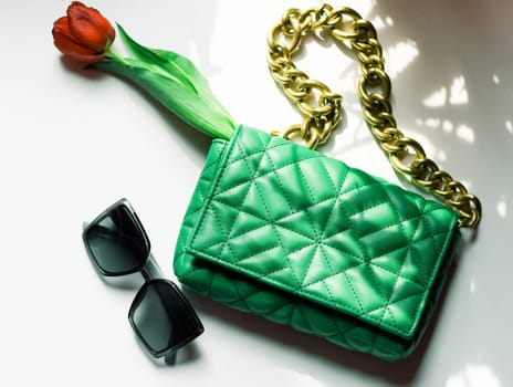 Stylish women's trendy green quilted bag with a large gold chain, a red tulip in it and sunglasses lie on a white table with shadows from the window, flat lay close-up. Fashionable accessories concept.