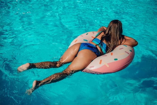 A woman is floating on a pink donut shaped float in a pool. The pool is blue and the woman is wearing a blue bikini