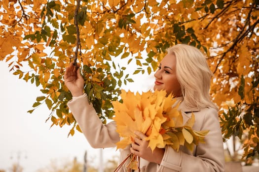 A blonde woman is holding a bunch of yellow leaves in her hand. She is standing in front of a tree with leaves that are changing colors. The scene has a peaceful and serene mood