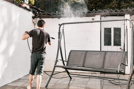 Portrait of a young brunette Caucasian guy in black clothes barefoot from the back washing a garden swing in the backyard of his house with a jet of water under pressure with a kercher, close-up view from the side.