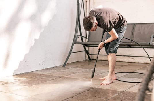 Portrait of a young brunette Caucasian guy in black clothes barefoot in a slanted position washing garden tiles in the backyard of his house with a jet of water under pressure with a kercher, close-up view from the side.