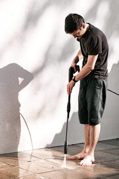 Portrait of a young brunette Caucasian guy in black clothes, barefoot, washing garden tiles with a jet of water under pressure with a kercher near a white wall at sunrise in the backyard of his house, close-up view from the side.