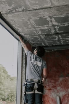 A young Caucasian man in a gray T-shirt with curly brown hair stands on a stepladder and cleans an old window opening with his hand to install a new frame, close-up view from below. The concept of installing windows, construction work, house renovation.