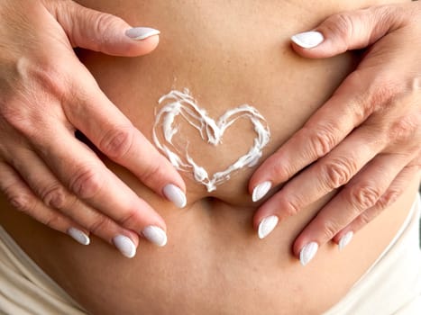 Close up of womans hands middle age with white nails applying cream in the shape of heart on her stomach. Skincare, self care, and personal hygiene with on love and body positivity. High quality photo