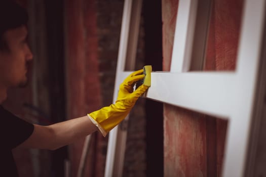 A young caucasian man in yellow gloves cleans a window frame with a sponge and soap, preparing them for installation, close-up side view. The concept of home renovation, installation of windows, construction work.