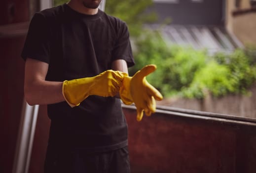 One young caucasian unrecognizable male builder in black uniform clothes pulling on yellow rubber gloves stretching his hand forward while standing near the window, close-up view from below. The concept of home renovation, construction work.