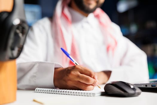Close-up shot of a notebook and pen being used by a Middle Eastern man in traditional clothing seated at a table. Selective focus on Muslim guy writing down research notes.