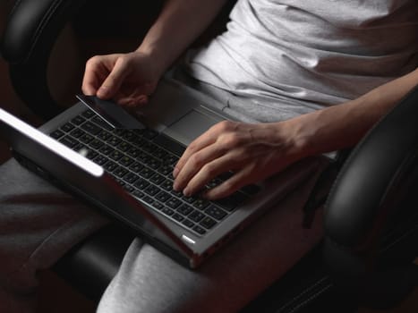 The hands of a young caucasian man in gray sweatpants and a t-shirt holds a bank card with one hand, and with the other he types his fingers on the keyboard while doing online shopping, sitting in a work chair in his room, side view close-up with depth of field. Online shopping concept, at home, style life, business, finance, gifts.