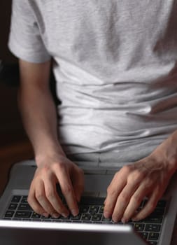 Hands of a young caucasian man in a gray t-shirt typing with his fingers on the keyboard doing online shopping, sitting in a work chair in his room, side view close-up with depth of field. Concept of online shopping, at home, lifestyle, business, finance, gifts.
