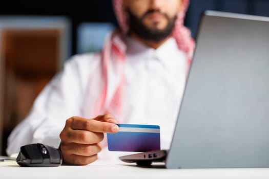 Man in Islamic attire sits at a modern office desk, browsing the internet on a laptop. He efficiently types and conducts research, using a credit card for secure online shopping and payment.