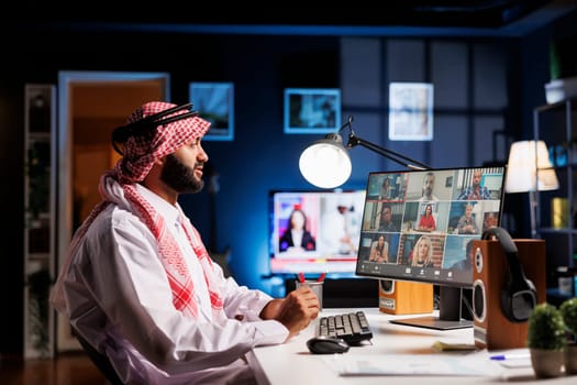 Side-view shot of a young man participating in an internet video conference with people. Muslim guy uses a desktop computer for study and communication while paying close attention to coworkers.