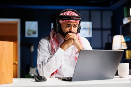 A focused man in traditional Arabic attire works diligently at his desk, utilizing wireless technology. He researches online, communicates, and engages in video conferencing on his personal computer.