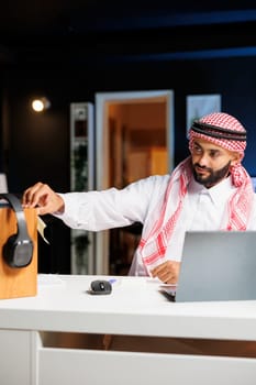 Middle Eastern man traditionally dressed is working on his laptop and taking notes while seated in his office. Muslim youngster placing sticky note on wooden board with headphones.