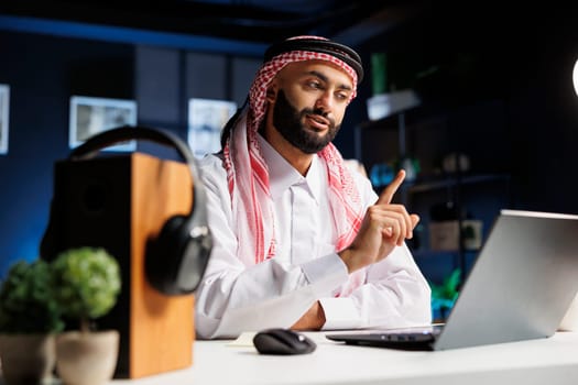 Close-up of an Arabian businessman dressed traditionally working intently at his contemporary desk, utilizing wireless technology and internet connection for research and video conferencing.