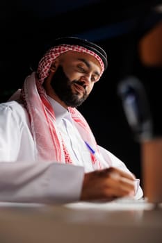 Detailed image of a concentrated Middle Eastern man in traditional clothing, working on a notepad at a table. Close-up of an Arab guy taking research notes at his home office with a pen.