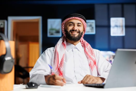 Portrait of a male Arab businessman looking at the camera, working at a desk with his digital laptop close by. Front-view shot of smiling Muslim guy writing on a notepad using a pen.