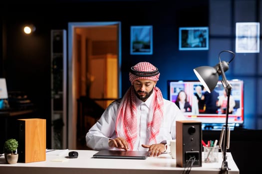 Middle Eastern guy dressed in Arabic attire sits at a table with his digital laptop, prepared for work. Committed Arab man plans to set up his personal computer to browse the internet.