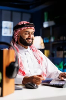 Side-view of Arab entrepreneur seated at a modern office desk engrossed in browsing the internet. A Muslim man in Islamic clothing, holding his debit card for online shopping on his personal computer.
