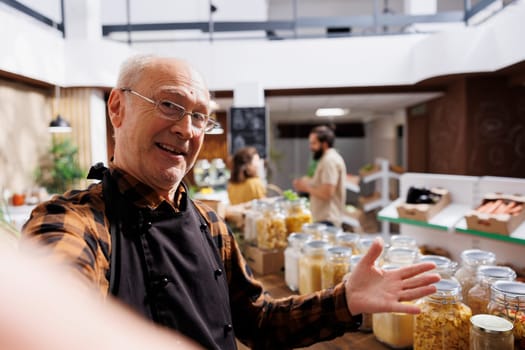 Older zero waste shop entrepreneur using selfie camera to show his natural pesticides free products online. Senescent supermarket owner promoting sustainable lifestyle, presenting his food stocks