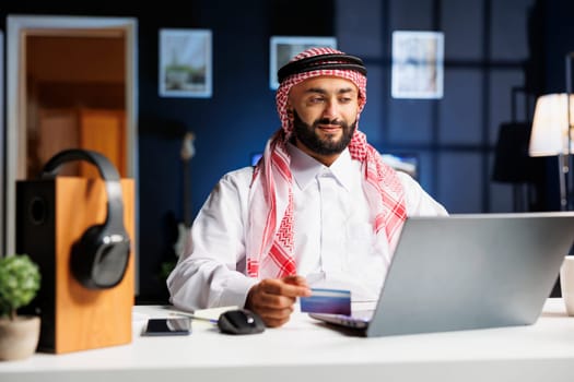 Arabic businessman in home office, efficiently browsing and typing on his laptop. He multitasks with online research, shopping, and payments using credit and debit cards.