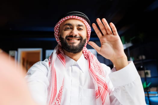POV of a cheerful Muslim guy dressed in traditional clothing, engaging in video call. Smiling Arab with thobe and headscarf grasping digital device and waving towards camera, doing online vlogging.