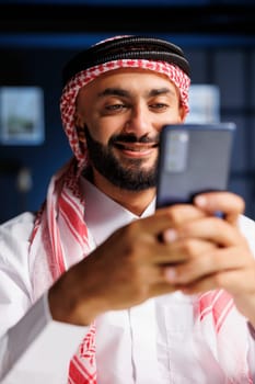 Detailed portrait shot of a smiling Muslim man using a smartphone for digital communication. Close-up view of an Arab entrepreneur typing and researching on his mobile device.