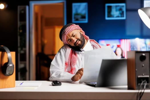 Professional Muslim man, dressed traditionally, works at his desk having a conversation on his smartphone while holding paperwork. Arab guy grasping research papers while talking on his cellphone.