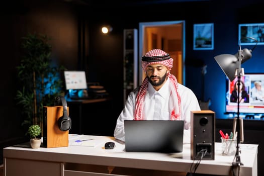 At modern workstation, confident Arab businessman types on his laptop. Muslim guy is browsing, conversing through email, and using WiFi connectivity in a calm and professional business environment.