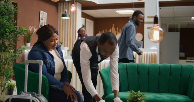 Bellhop providing coffee cup for client sitting on lounge area couch at hotel, staff offering great luxury services with drink from bar. African american tourist receiving beverage in lobby.