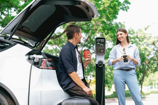 Lovely young couple drinking coffee while recharging battery for electric car during road trip travel EV car in natural forest or national park. Eco friendly travel during vacation and holiday. Exalt