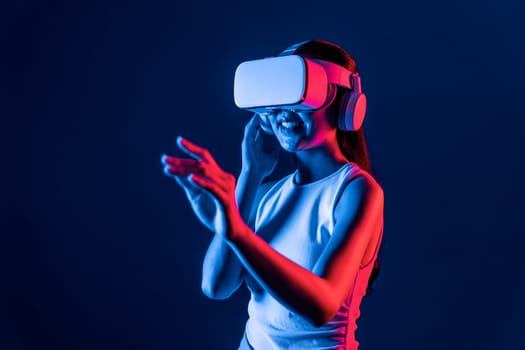 Smart female standing with surrounded by cyberpunk neon light wear VR headset connecting metaverse, futuristic cyberspace community technology. Woman using hand touching virtual object. Hallucination.