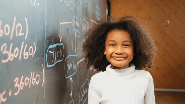 Site view of young african girl looking at camera while standing at blackboard with engineering prompt. Student smiling at camera system code written behind in STEM technology classroom. Erudition.