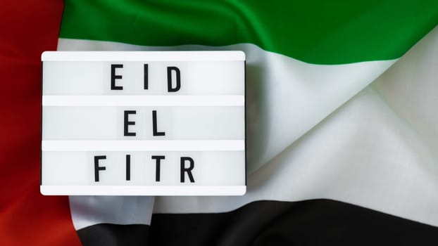 EID EL FITR - Eid Mubarak - Happy Holidays text frame on United Arab Emirates waving flag made from silk material. Public holiday celebration background. The National Flag of UAE. Patriotism Commemoration Day Muslim Ramadan Blessed Holy Month concept