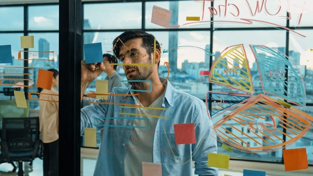 Professional businessman uses sticky notes at glass wall with graph to brainstorming idea while multicultural businesspeople discussing about marketing ideas at office with city view. Tracery