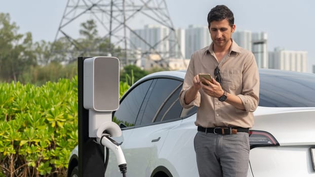 Man pay for electricity with smartphone while recharge EV car battery at charging station connected to power grid tower electrical as electrical industry for eco friendly car utilization.Expedient