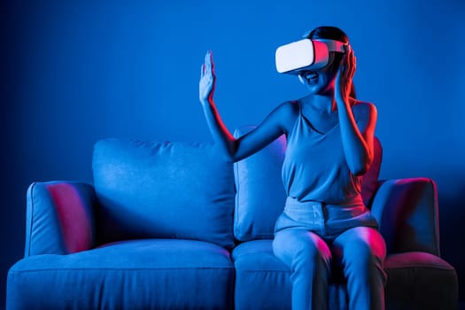 Smart female sitting on sofa wearing VR headset connecting metaverse, future cyberspace community technology. Elegant woman enjoy gesticulate interacting another users in virtual world. Hallucination.