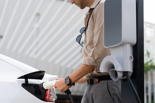 Young man put EV charger to recharge electric car's battery from charging station in city commercial parking lot. Rechargeable EV car for sustainable environmental friendly urban travel. Expedient