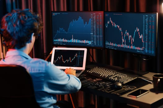 Young investor stock trader searching on laptop comparing with dynamic exchange rate screens of current market graph online in real time at neon light modern office, analyzing financial data. Gusher.