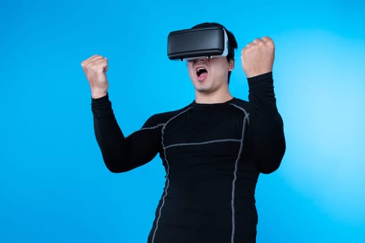Smart gaming player wearing VR glasses raising fist up isolated blue background screen connecting digital futuristic technology virtual reality at metaverse world playing hologram arena. Contrivance.