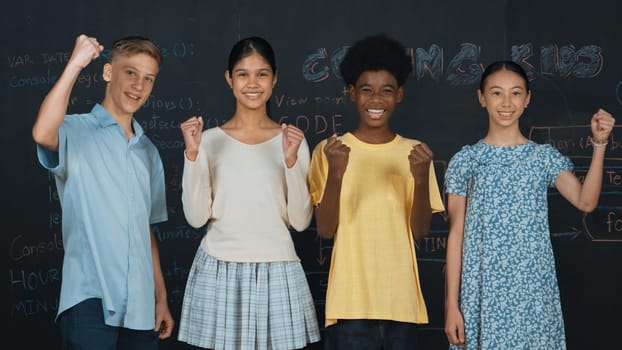 Diverse academic student cheering and celebrate successful project while raised hand. Group of multicultural children standing at blackboard with engineering code while looking at camera. Edification.