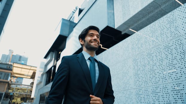 Smart business man wearing suit and smiling while walking at street near white modern architectural building. Attractive handsome manager looking skyscraper in urban city. Copy space. Exultant.