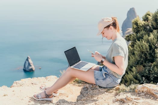 Digital nomad, woman in the hat, a business woman with a laptop sits on the rocks by the sea during sunset, makes a business transaction online from a distance. Freelance, remote work on vacation.