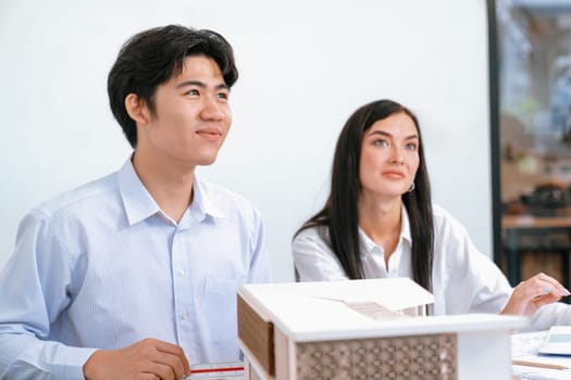 Professional male asian architect using ruler to measure house model length while young beautiful caucasian colleague using laptop to analyzed data on meeting table with house model. Immaculate.