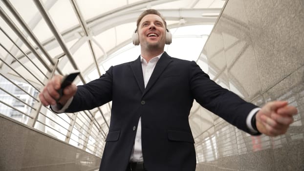 Business man dance to relaxing music from headphone at modern corridor. Cheerful project manager wear headset and moving gesture to celebrate successful song with lively mood in urban city. Urbane.