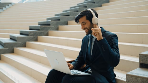 Confident smart business man celebrate successful business idea while listen music. Happy manager project working by using laptop while wearing headphone and suit. Investor sitting at stair. Exultant.