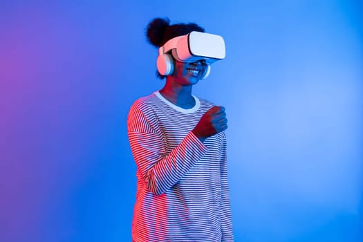 Smiling young African woman wearing VR headset smelling good through metaverse flowers filed world at vibrant neon pink blue studio lighting background technology learning 3D cyberspace. Contrivance.