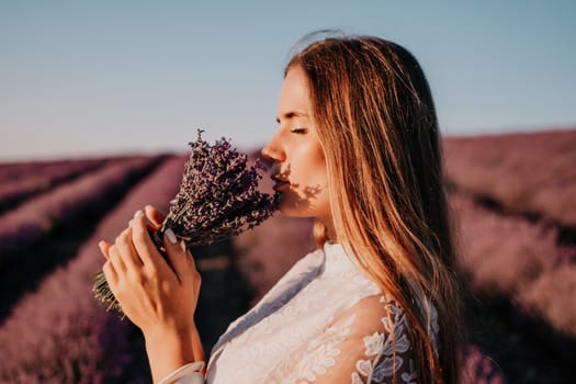 Close up portrait of young beautiful woman in a white dress and a hat is walking in the lavender field and smelling lavender bouquet.