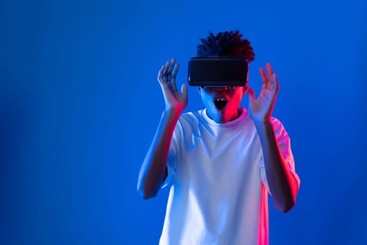 Surprised young African American looking through VR holding interesting object hologram on pink blue neon wall at metaverse world connecting digital futuristic technology virtual reality. Contrivance.