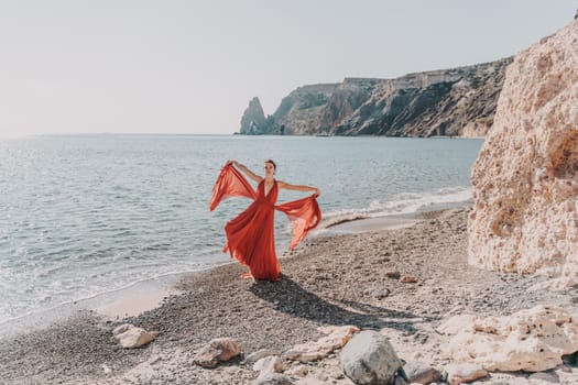 Woman red dress sea. Female dancer in a long red dress posing on a beach with rocks on sunny day. Girl on the nature on blue sky background