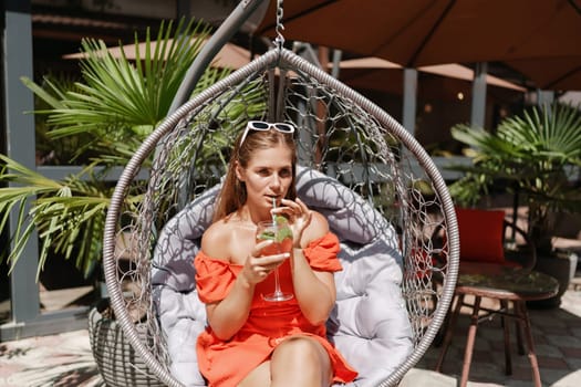A woman in an orange dress is sitting in a hanging chair. She is holding a wine glass and smiling. The scene takes place in a restaurant with several chairs and tables